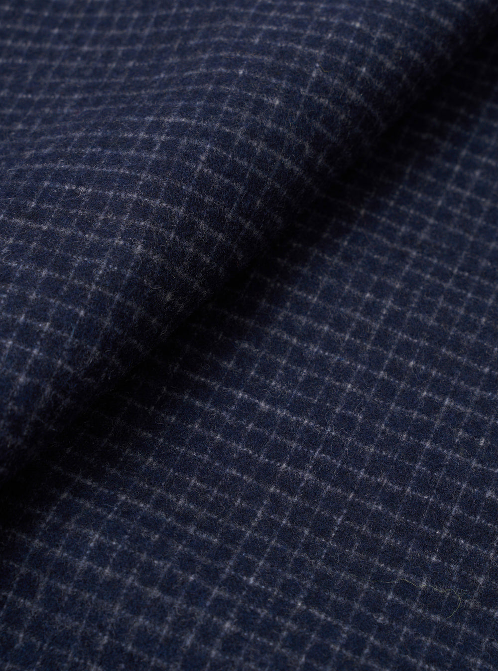 Close-up fabric shot of the navy check wool mix used in the Bobby Bakers Overshirt. A fine grey windowpane check runs across a navy ground, with a soft, lightly hairy wool texture visible across the draped and folded cloth.