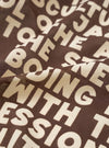 A close-up fabric swatch view of the Brown Jazz/Beatniks Print Neckerchief on a solid backdrop, showing the detailed text graphic design and the texture of the soft cotton material.