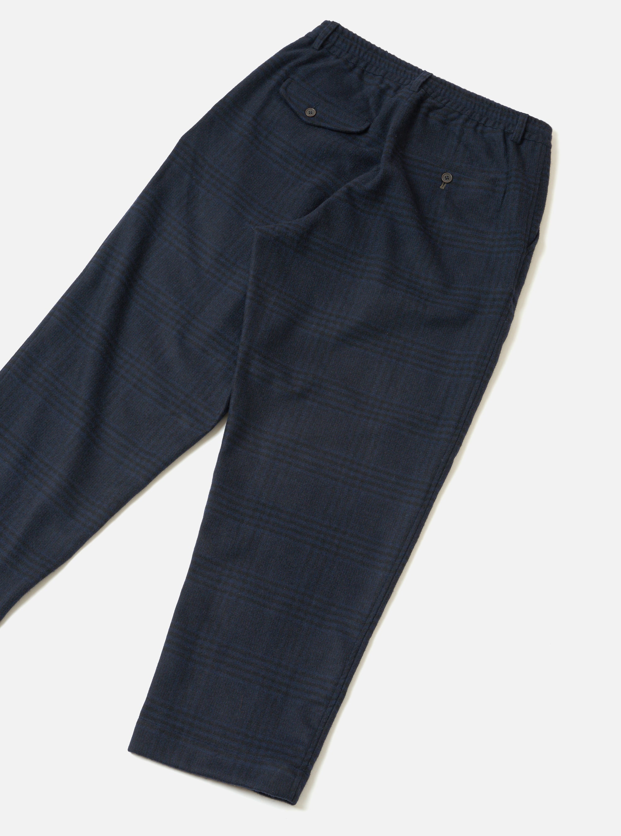 Navy wool-mix oxford trousers laid flat on a light grey seamless surface. The fabric features a subtle glen check pattern in navy and dark green. The elasticated waistband is visible at the top, with a buttoned flap pocket on the left hip and a small button-fastened welt pocket on the right rear. Tapered leg with a plain hem.