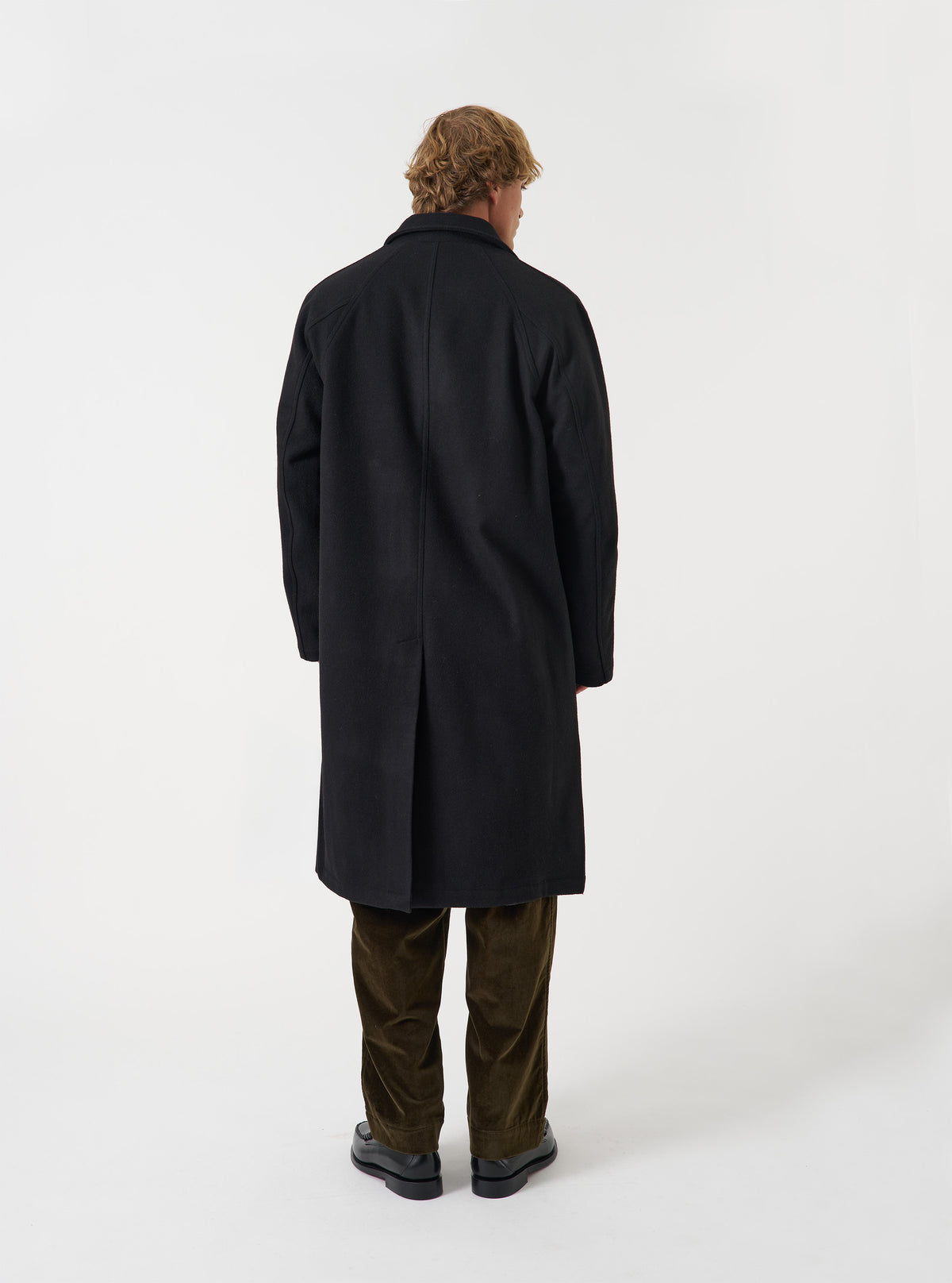 An alternative view of a model wearing the Black Mowbray Mac Coat on a neutral backdrop, highlighting the longline silhouette, classic collar detail, and tailored dark grey-black fit.