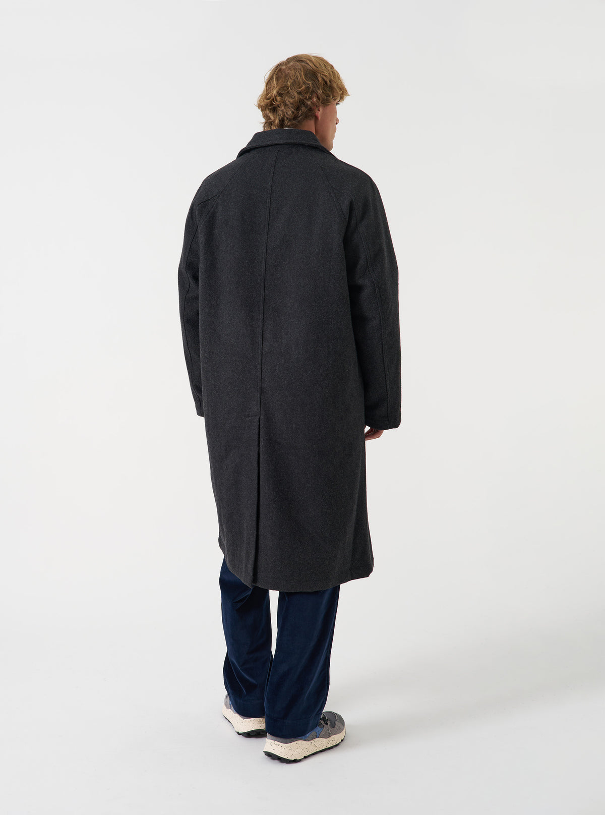 A model viewed from a full-length perspective wearing the Grey Marl Mowbray Mac Coat set against a neutral studio backdrop, highlighting its clean silhouette, back vent, and relaxed fit.