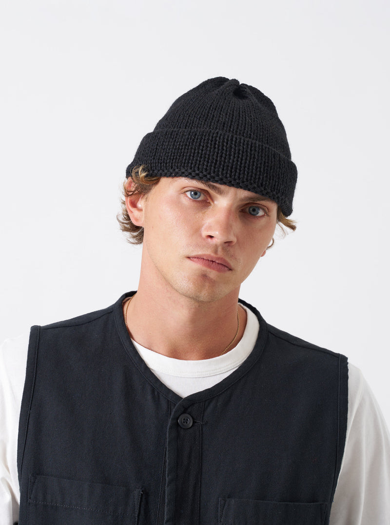 A model wearing the Black British Wool Short Watch Cap against a clean studio background. The short beanie sits close above the ears and features a turned-up cuff and a pinched crown profile.