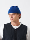 A model wearing the Bright Navy British Wool Short Watch Cap against a neutral studio backdrop. The close-fitting knitted hat features a folded brim and sits neatly above the ears of the model.