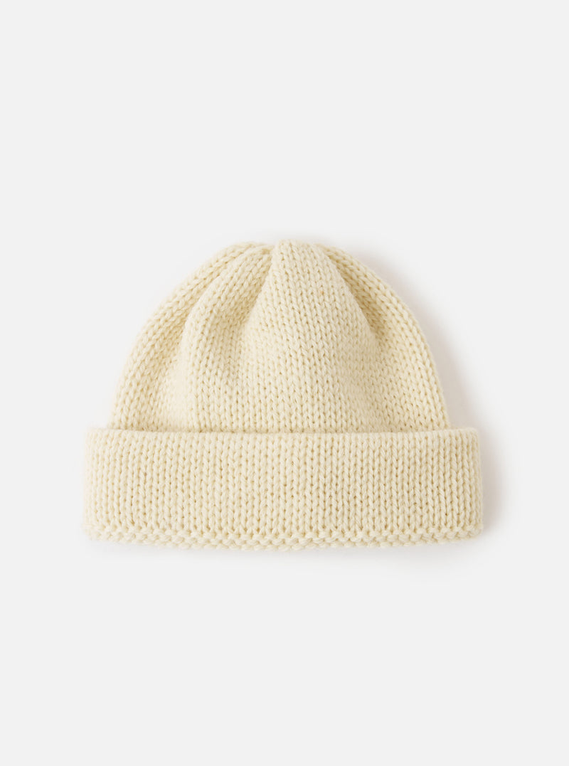 The Ecru British Wool Short Watch Cap shown flat against a light studio backdrop. Knitted from 100% British wool in an off-white shade, the hat features a turned-up cuff and ribbed texture.
