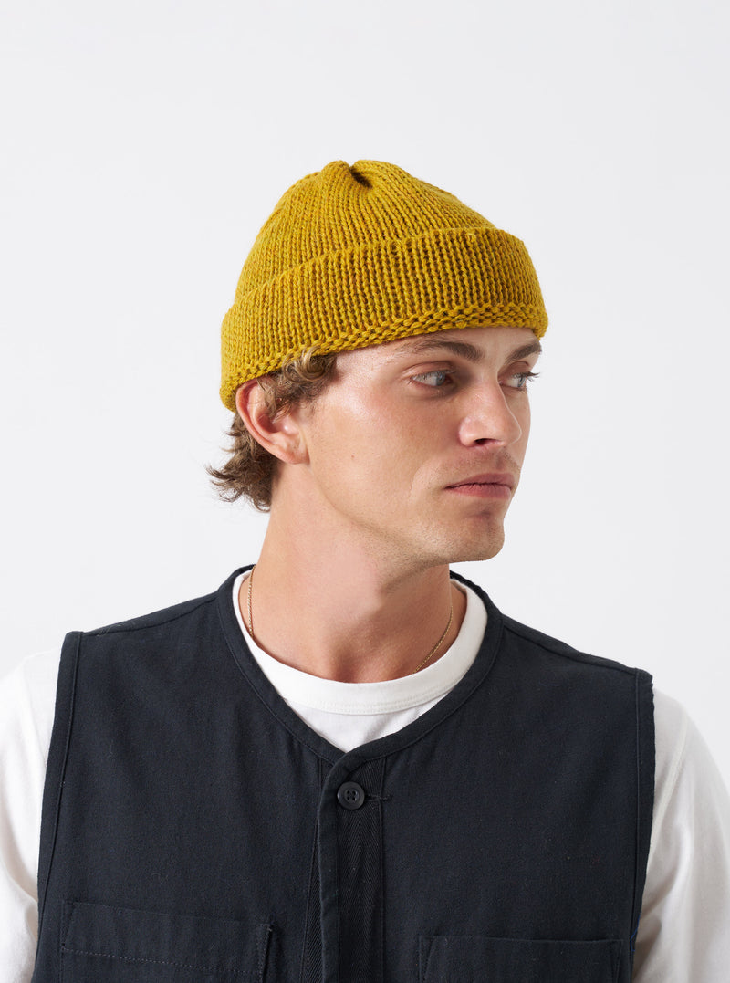 A model wearing the Mustard British Wool Short Watch Cap against a neutral backdrop. The bright yellow-ochre beanie features a short fit that sits above the ears with a folded cuff.
