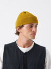 A model wearing the Mustard British Wool Short Watch Cap against a neutral backdrop. The bright yellow-ochre beanie features a short fit that sits above the ears with a folded cuff.