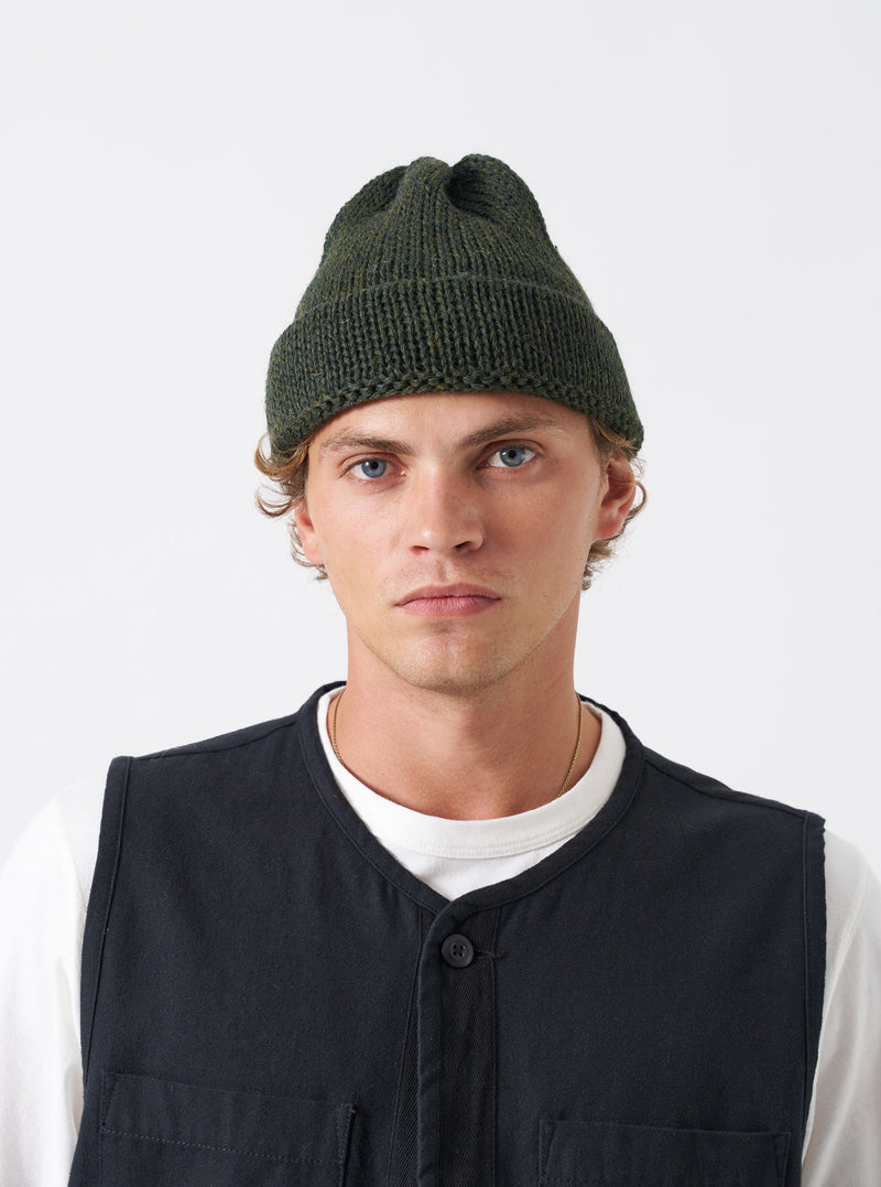 A model wearing the Olive British Wool Short Watch Cap against a plain studio background. The knitted beanie hat features a folded brim, rich olive green wool texture, and a classic ribbed construction.