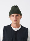 A model wearing the Olive British Wool Short Watch Cap against a plain studio background. The knitted beanie hat features a folded brim, rich olive green wool texture, and a classic ribbed construction.