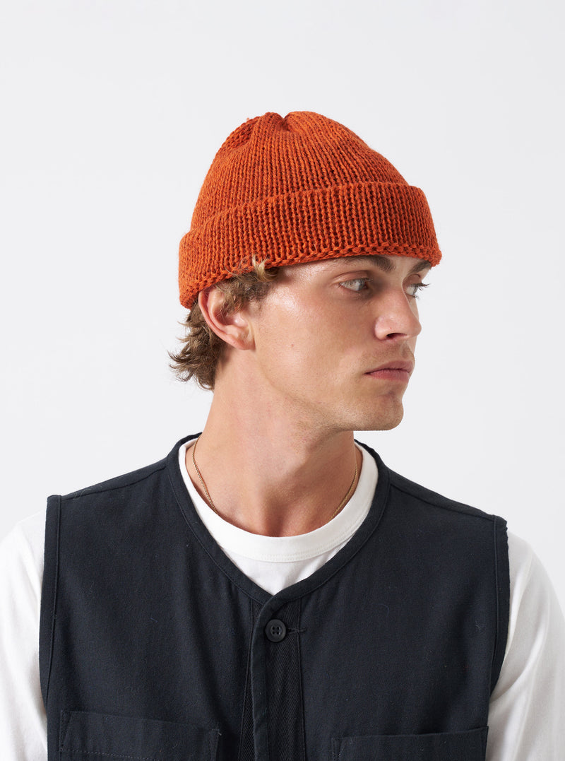 A model wearing the bright Orange British Wool Short Watch Cap against a clean studio background. The short knitted beanie hat fits neatly above the ears and showcases a ribbed wool texture.