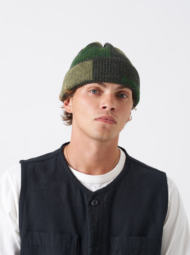 A model wearing the Patched Olive British Wool Short Watch Cap against a neutral studio background. The olive green knitted beanie fits neatly above the ears and features a subtle patch accent.