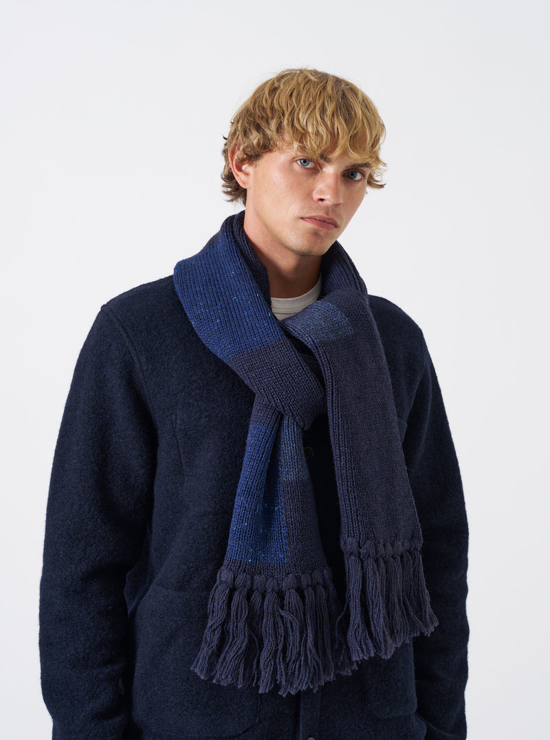 A model wearing the Navy British Wool Scarf wrapped styled around the neck against a plain background. The dark navy wool scarf displays its full length, soft drape, and textured ribbed knit ends.