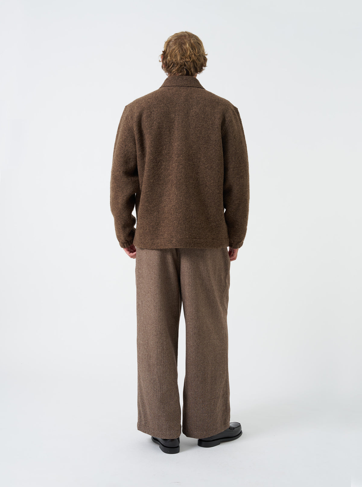 A male model is shown from the side wearing the Brown Marl Wool Fleece Rams Jacket in a studio setting. The shot demonstrates the relaxed silhouette, drop shoulder detail, and comfortable fit of the garment.