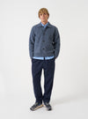 A male model wears the Slate Blue Wool Fleece Rams Jacket against a plain background. The jacket is styled open over a t-shirt, showing its casual boxy fit, dropped shoulders, and prominent patch pockets.