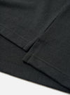 Close-up flat lay detail of the black recycled wool mix long-sleeve pocket tee. The shot focuses on the chest patch pocket and sleeve cuff, showing the folded ribbed cuff edge with visible topstitching and a zigzag overlock seam. The fine-knit jersey fabric has a subtle heathered texture. White surface background.