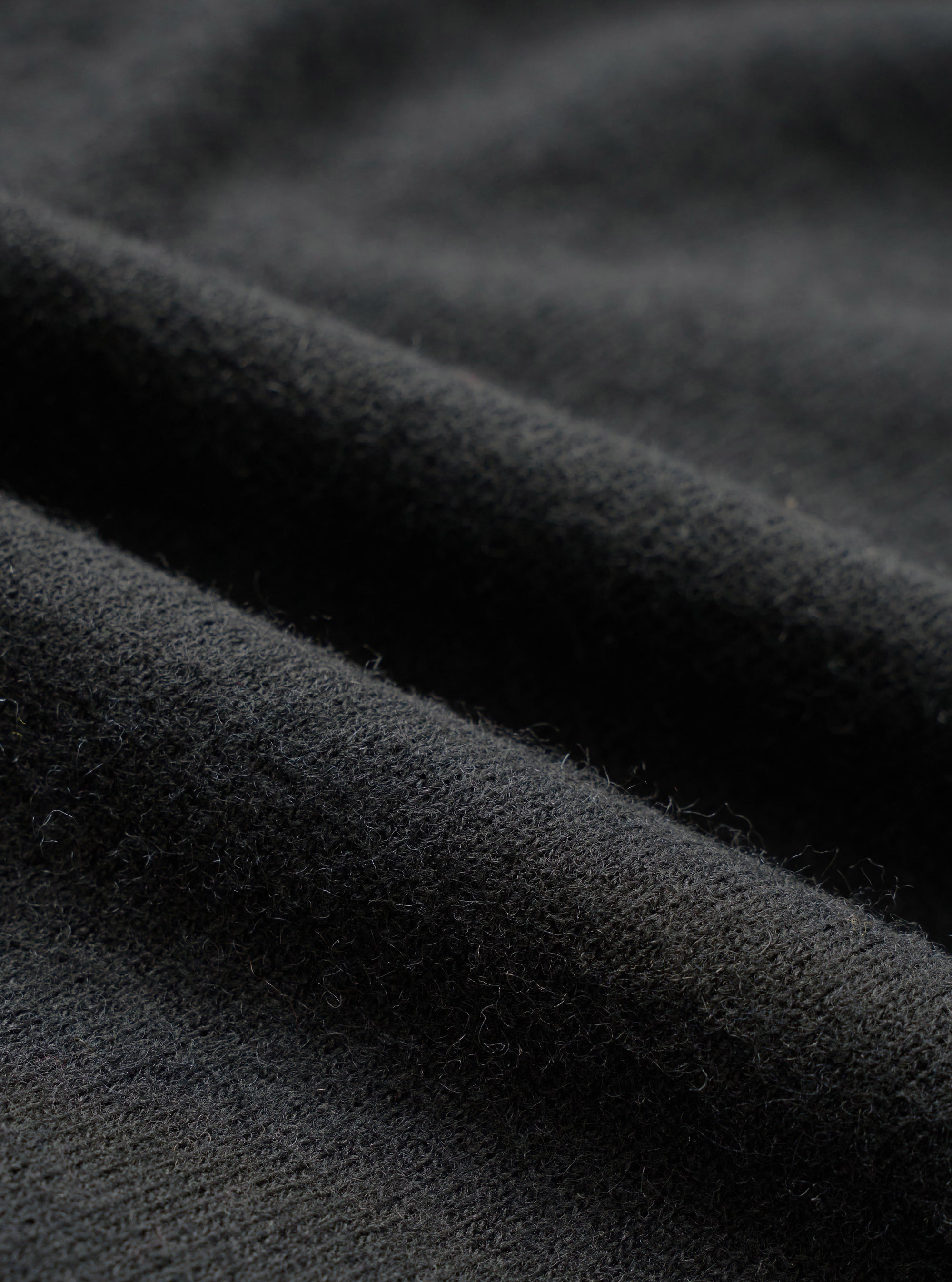Close-up detail shot of the black recycled wool mix fabric, showing a soft, lightly brushed texture with fine fibres visible on the surface. The material has a dense, matte finish with gentle folds catching the light to reveal subtle tonal variation across the dark charcoal-black cloth.