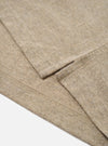 Close-up detail of a dark sand recycled wool mix long-sleeve pocket tee, flat on a white surface. The shot focuses on the curved hem with visible overlock stitching and a small side seam vent. The marl-textured fabric shows a heathered oatmeal tone with flecks of darker fibre throughout.