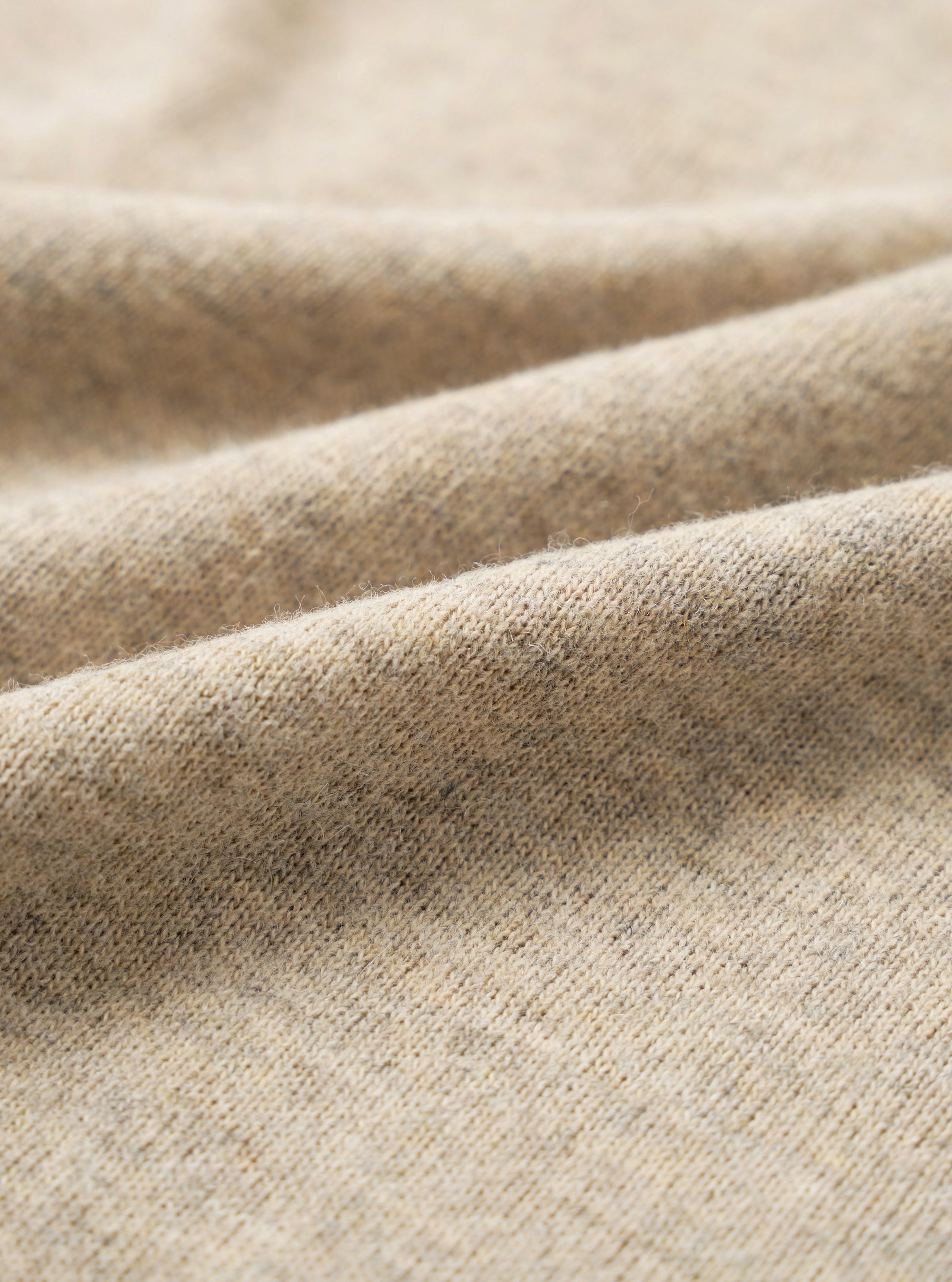 Close-up of dark sand recycled wool mix fabric, showing a fine knit texture with a soft, slightly haired surface. The oatmeal-toned yarn has subtle flecks of grey and brown throughout. Gentle folds run diagonally across the frame, highlighting the fabric's drape and weight.