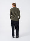 Studio shot of a model wearing the Olive Recycled Wool Mix SJ L/S Pocket Tee, showing the side profile and relaxed fit of the long-sleeve shirt with its crew collar and self-fabric cuffed sleeves.