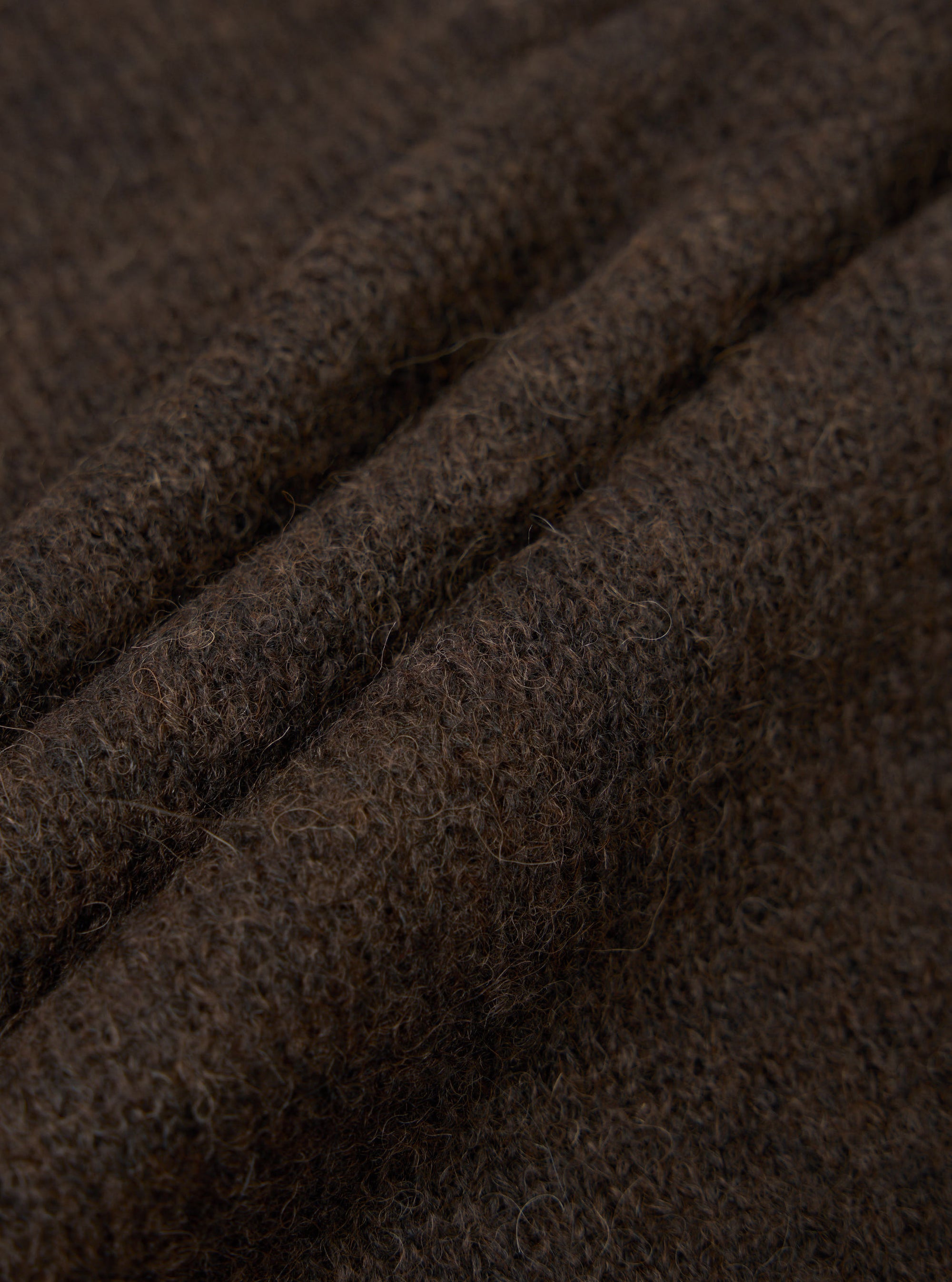 Close-up detail shot of the brown alpaca knit fabric, showing a hairy texture with visible individual fibres. Three soft ridges of folded knit run diagonally across the frame, highlighting the fabric's lofty, textured hand. Dark brown melange colouring throughout.