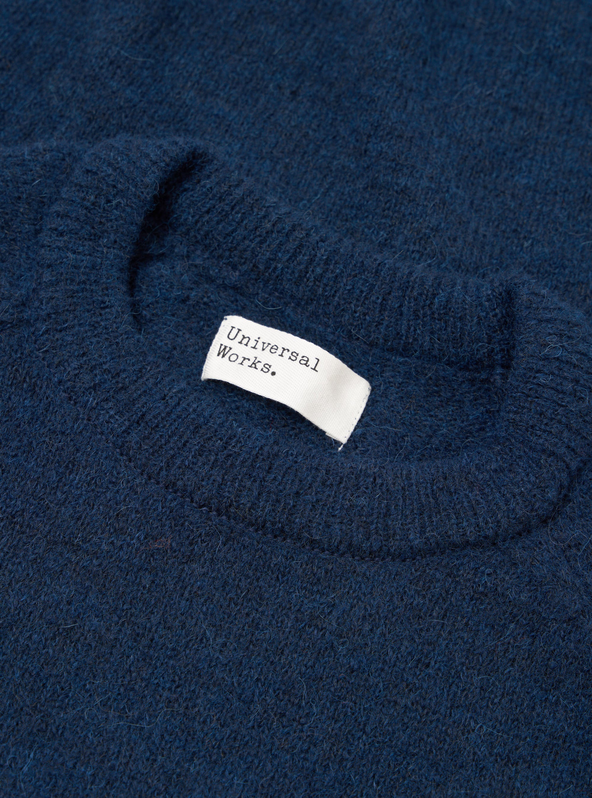 Close-up flat lay of the crew neck collar on a navy alpaca knit jumper. The ribbed neckline has a soft, fluffy texture. A white woven Universal Works label is visible inside the collar. The fabric shows a marl-flecked deep navy tone throughout.