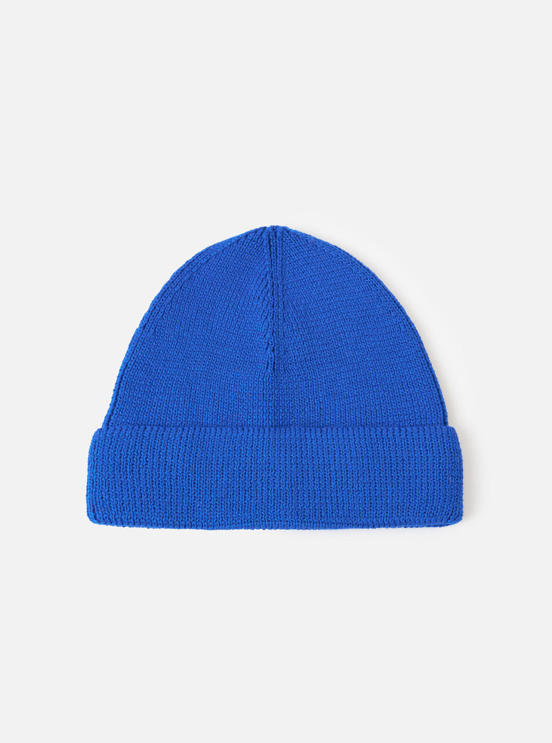 A Blue Merino Knit Watch Cap displayed flat against a plain studio background. The classic knitted beanie features a soft ribbed texture, a turn-up cuff, and a comfortable stretchy fit in blue merino wool.