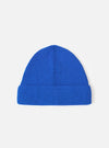 A Blue Merino Knit Watch Cap displayed flat against a plain studio background. The classic knitted beanie features a soft ribbed texture, a turn-up cuff, and a comfortable stretchy fit in blue merino wool.