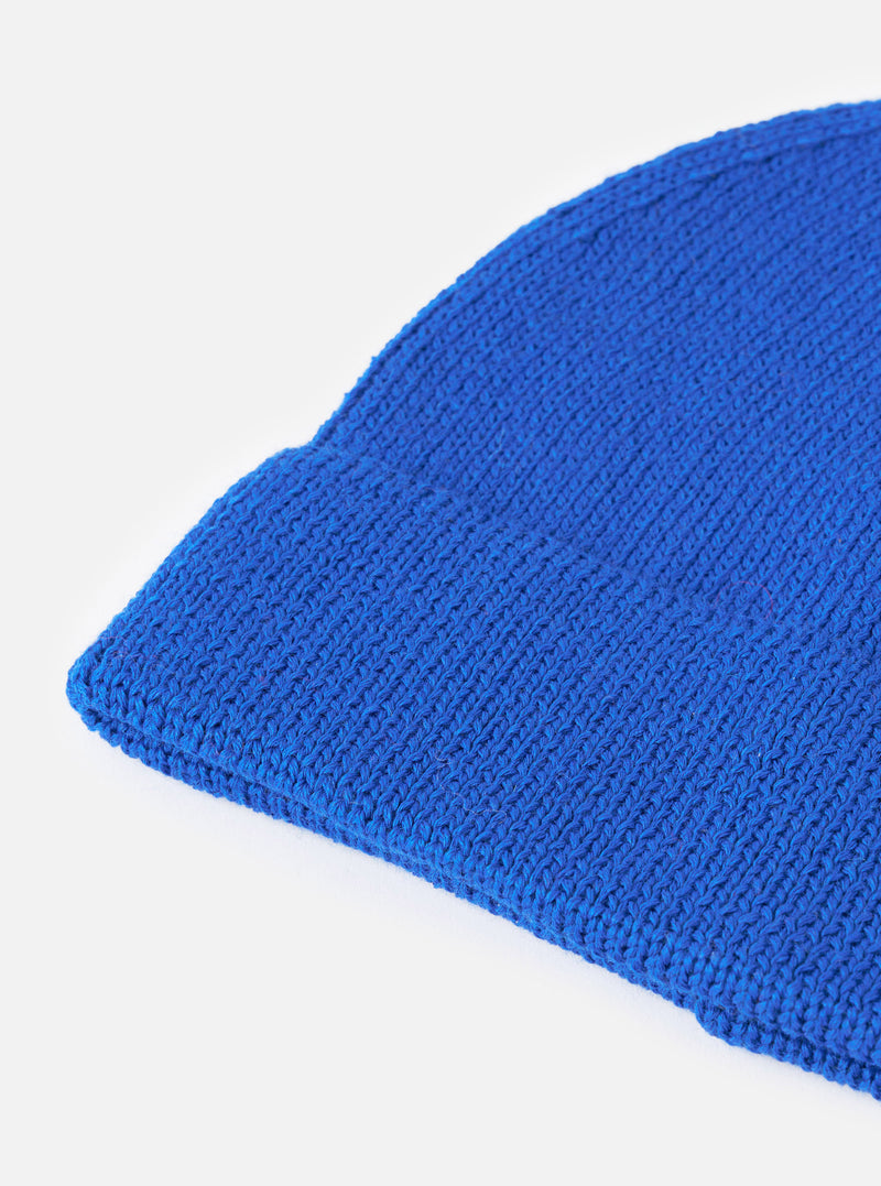 An angled view of the Blue Merino Knit Watch Cap on a plain neutral background, highlighting the full shape of the ribbed beanie, pinched crown construction, and rolled cuff detail in vibrant blue.
