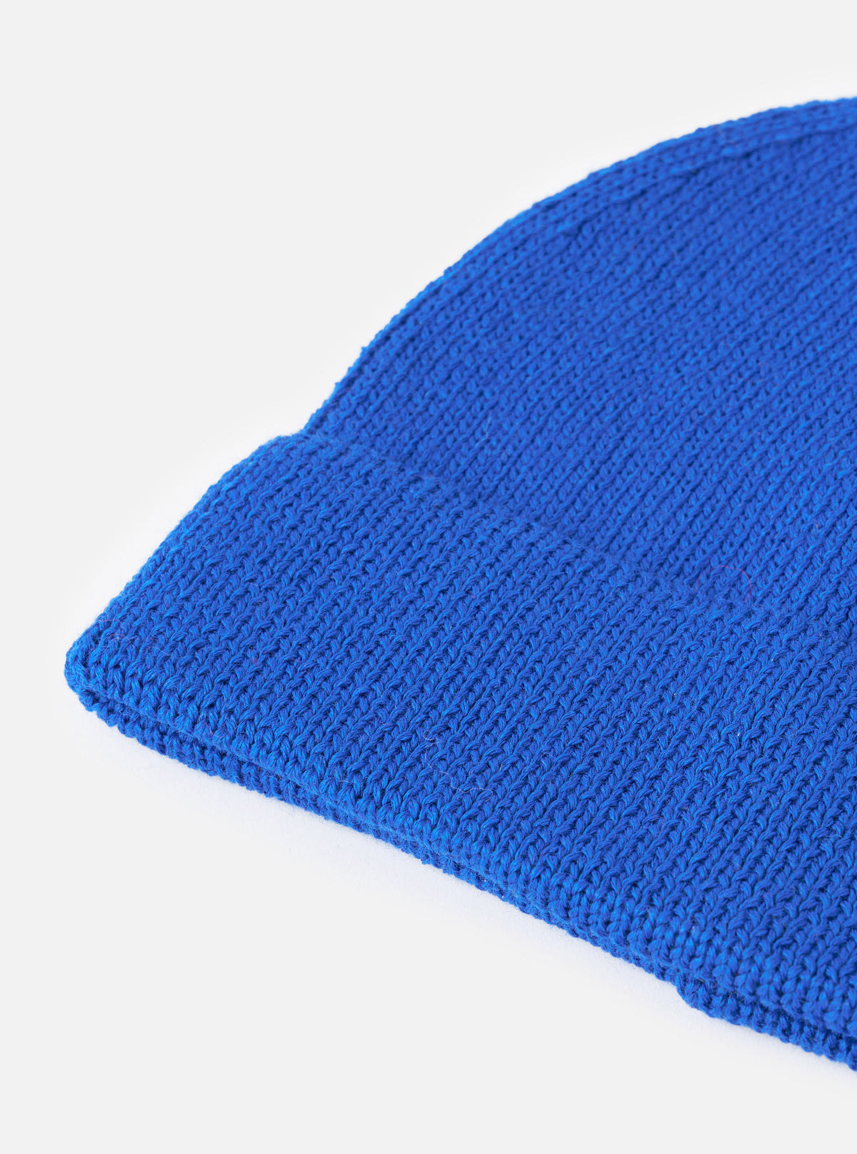 An angled view of the Blue Merino Knit Watch Cap on a plain neutral background, highlighting the full shape of the ribbed beanie, pinched crown construction, and rolled cuff detail in vibrant blue.