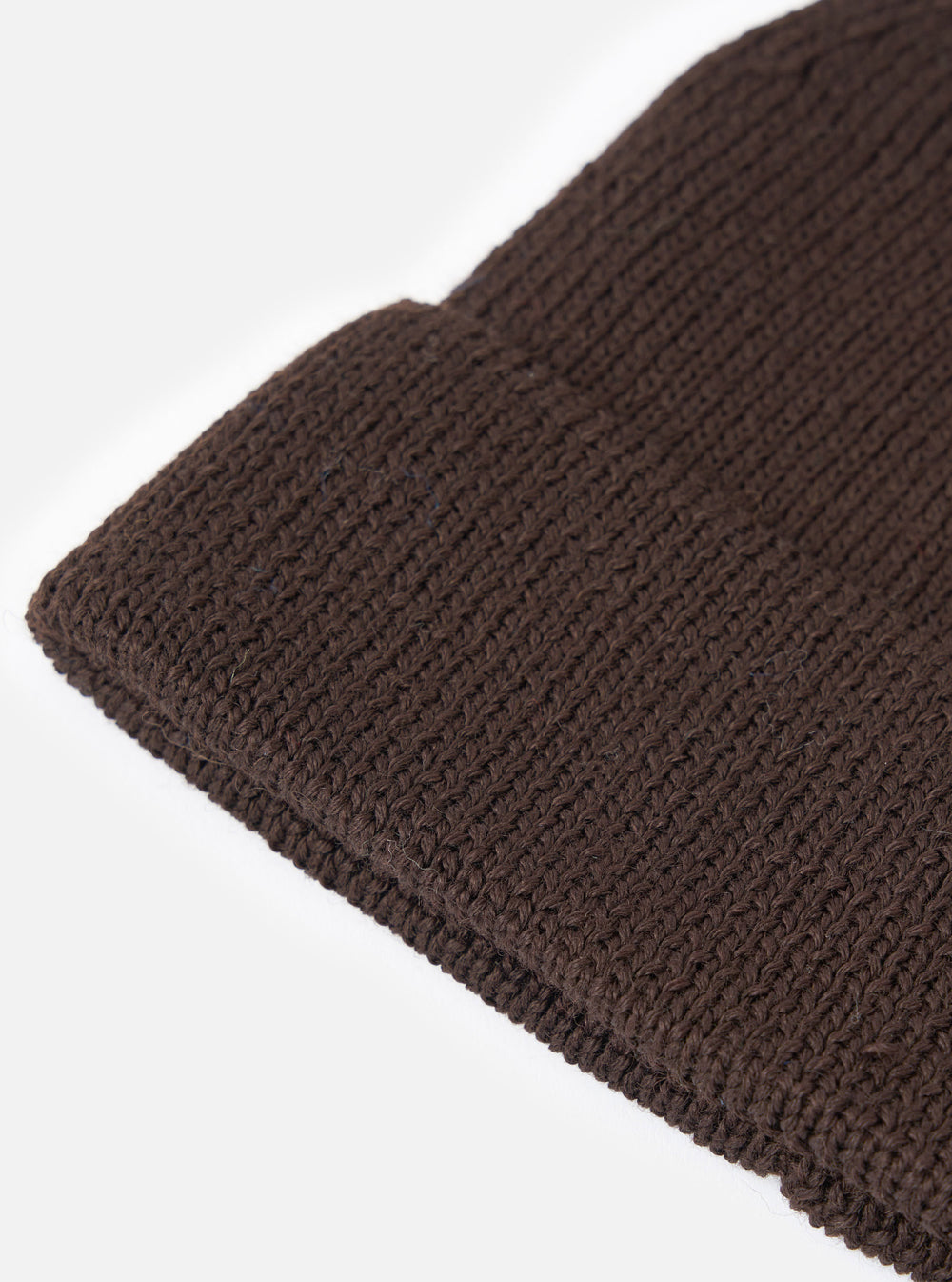 An angled view of the Brown Merino Knit Watch Cap on a plain neutral background, highlighting the full shape of the ribbed beanie, pinched crown construction, and rolled cuff detail in rich brown wool.