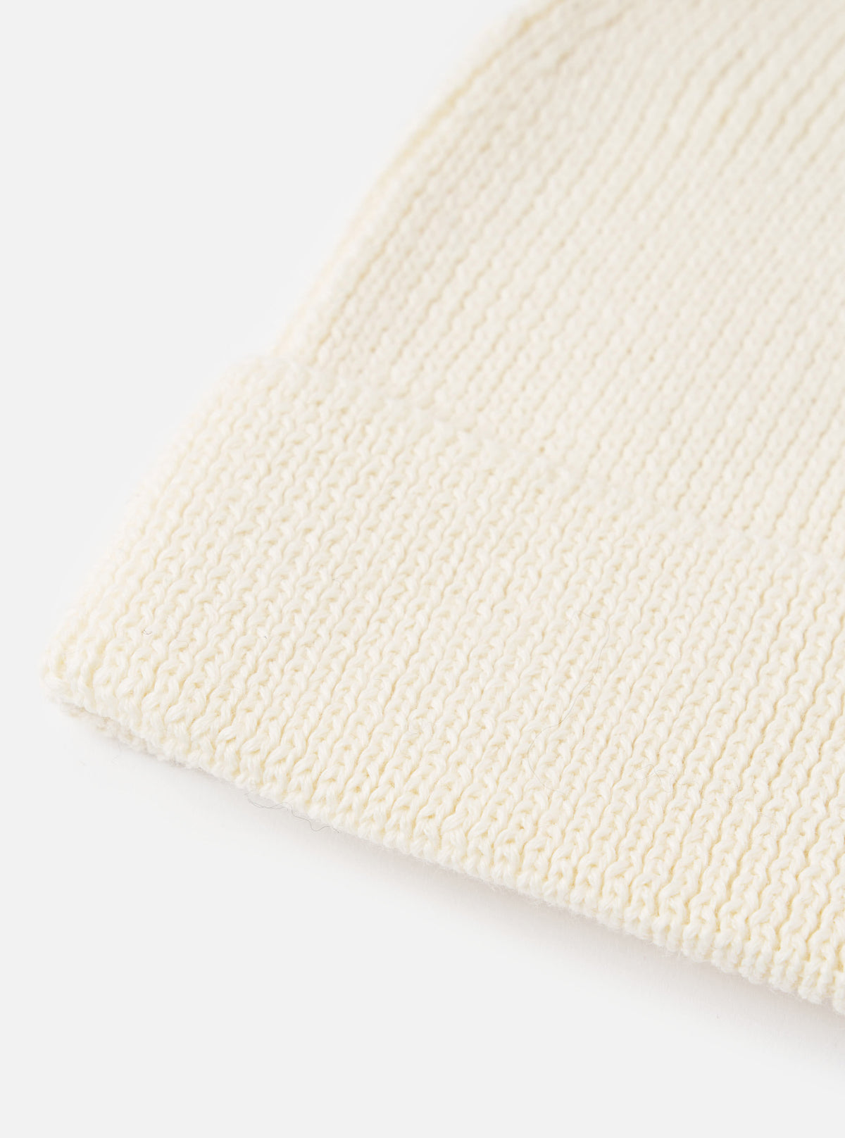An angled view of the Ecru Merino Knit Watch Cap on a plain neutral background, highlighting the full shape of the ribbed beanie, pinched crown construction, and rolled cuff detail in off-white ecru wool.