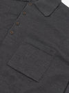 Close-up flat lay of a charcoal merino long-sleeve knit polo shirt on a white surface. The fine-knit fabric has a smooth, dense texture. Visible details include a spread collar, a four-button placket with dark tonal buttons, and a large patch chest pocket with a horizontal seam detail at the base.