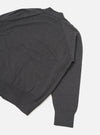 Flat lay of a charcoal merino loose cardigan on a white studio backdrop, shot from above at an angle. The fine-knit body shows a mock neck or collar at the top, a ribbed hem, and one full sleeve with a ribbed cuff. A seam detail runs along the sleeve join.