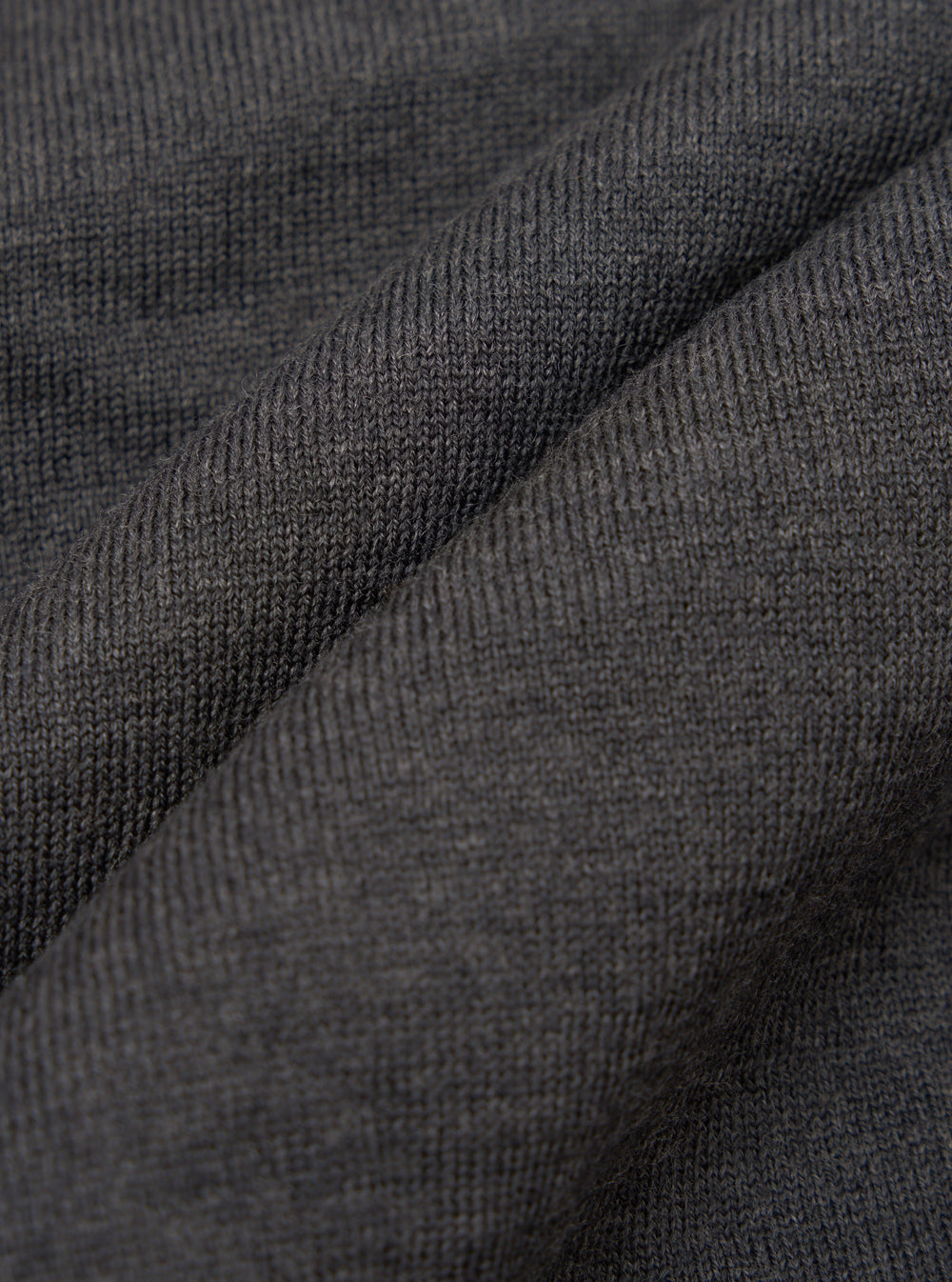 Close-up of charcoal merino knit fabric, showing a fine, even stocking stitch with a soft, slightly heathered texture. The fabric is gathered into soft folds, highlighting the drape and weight of the knit. No background is visible.