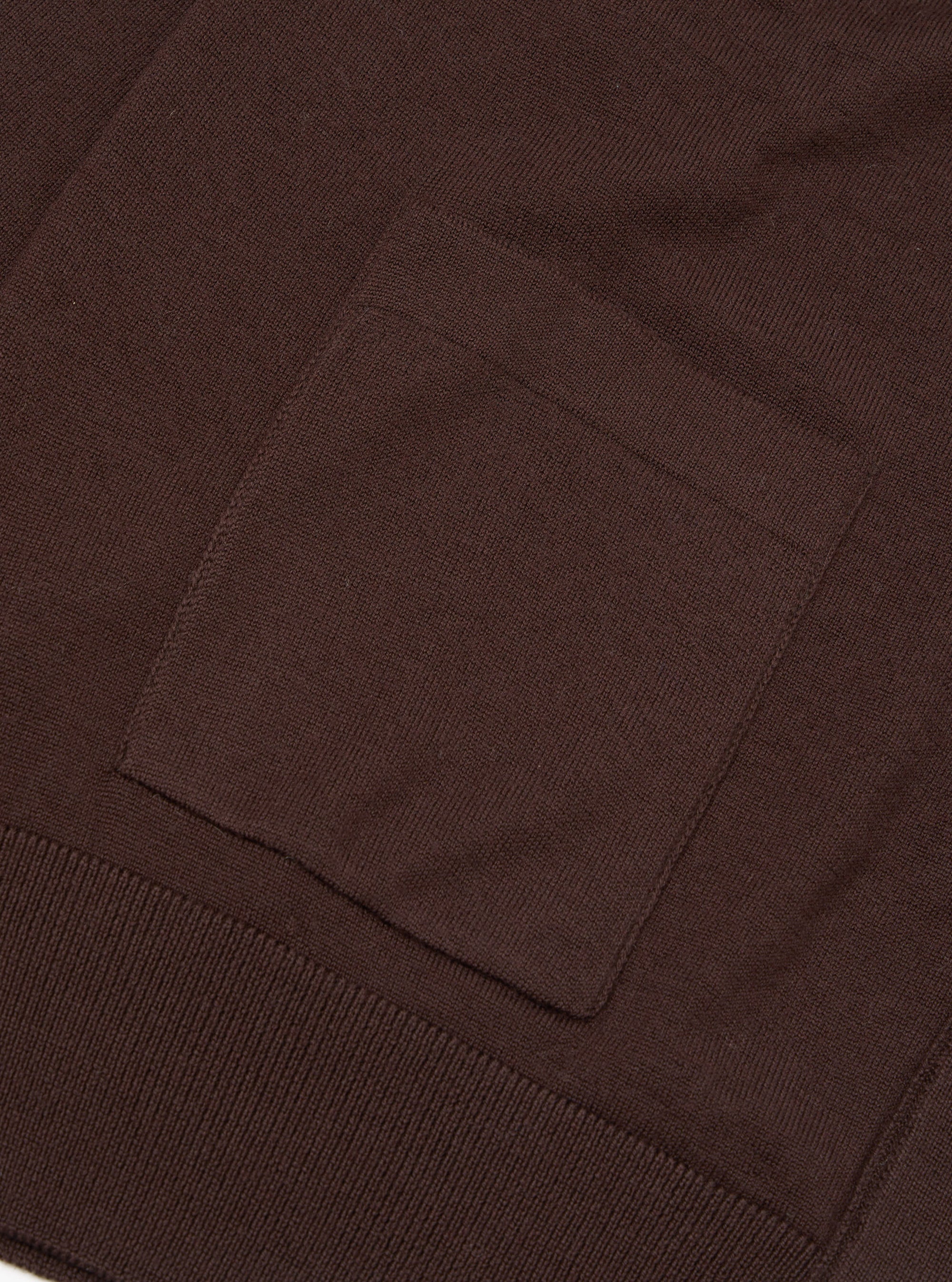 Close-up flat lay of the lower section of an espresso brown merino cardigan. A square patch pocket sits above a ribbed hem band. The fine knit texture is visible across the body. Plain white studio backdrop.