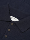 Close-up flat lay of a navy Shetland knit sweater showing the collar and button placket. The textured knit fabric is visible throughout. A white Universal Works woven label sits inside the collar. Three dark horn-effect buttons are visible on the placket. Shot on a plain white studio backdrop.