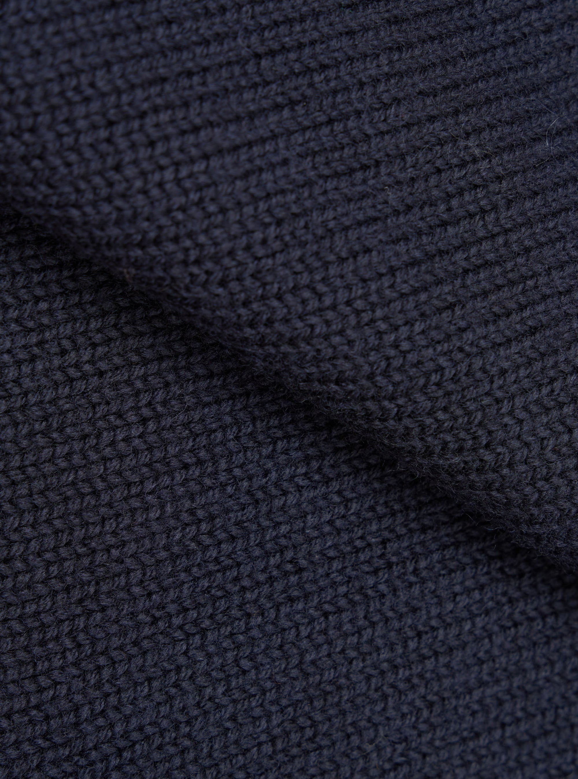 Close-up detail shot of the navy Shetland Wild Curtis sweater fabric. The chunky knit texture is shown in high detail, revealing a moss or seed stitch construction with a ribbed section visible at the top. The wool yarn is a deep navy, shot against no background.
