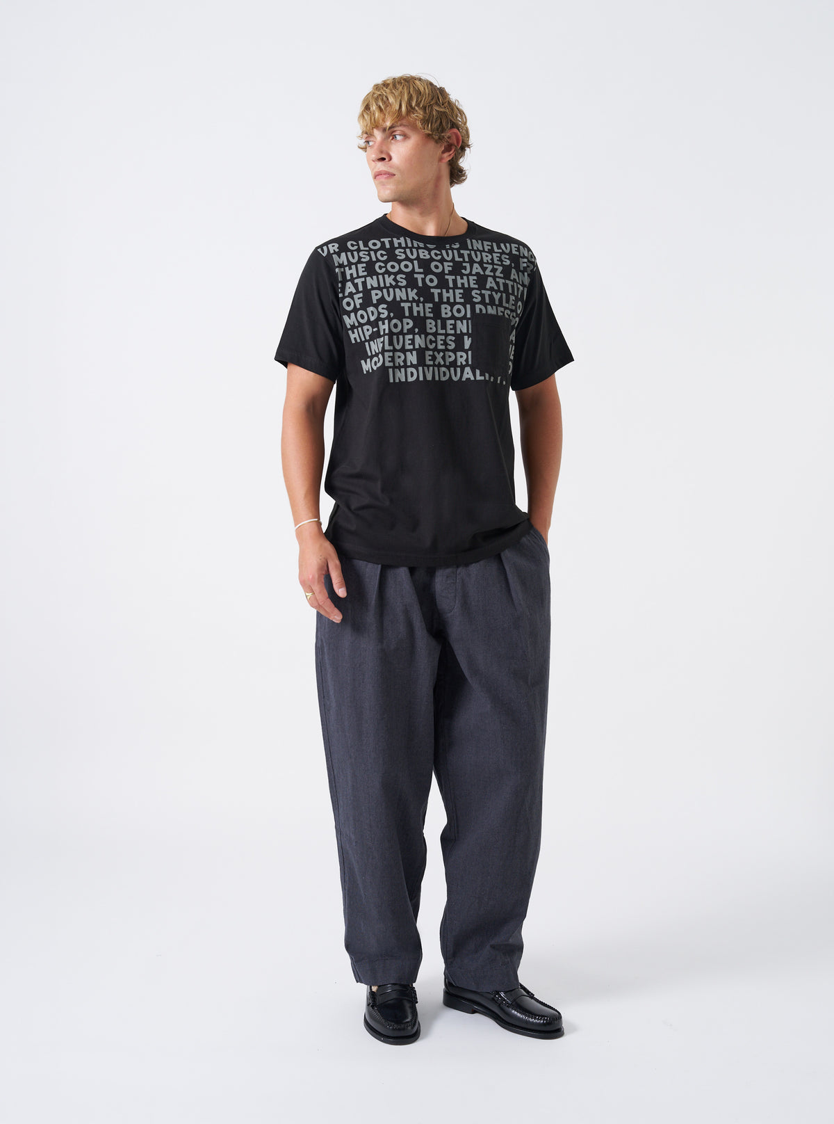 A male model standing against a plain studio background wearing the Grey Liverpool Check Oxford Pant paired with casual footwear, demonstrating the relaxed loose fit, wide leg silhouette, and drape of the grey check trousers.