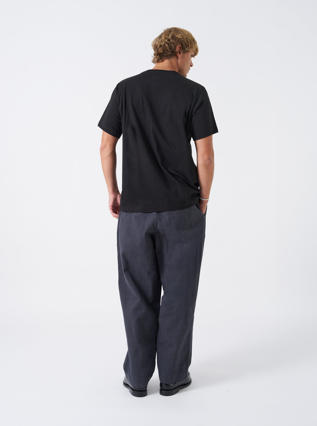 A model wearing the Grey Liverpool Check Oxford Pant, shown from the side against a neutral white backdrop. The photo highlights the comfortable fit, pleated waistband, side pockets, and full length drape of the checked trousers.