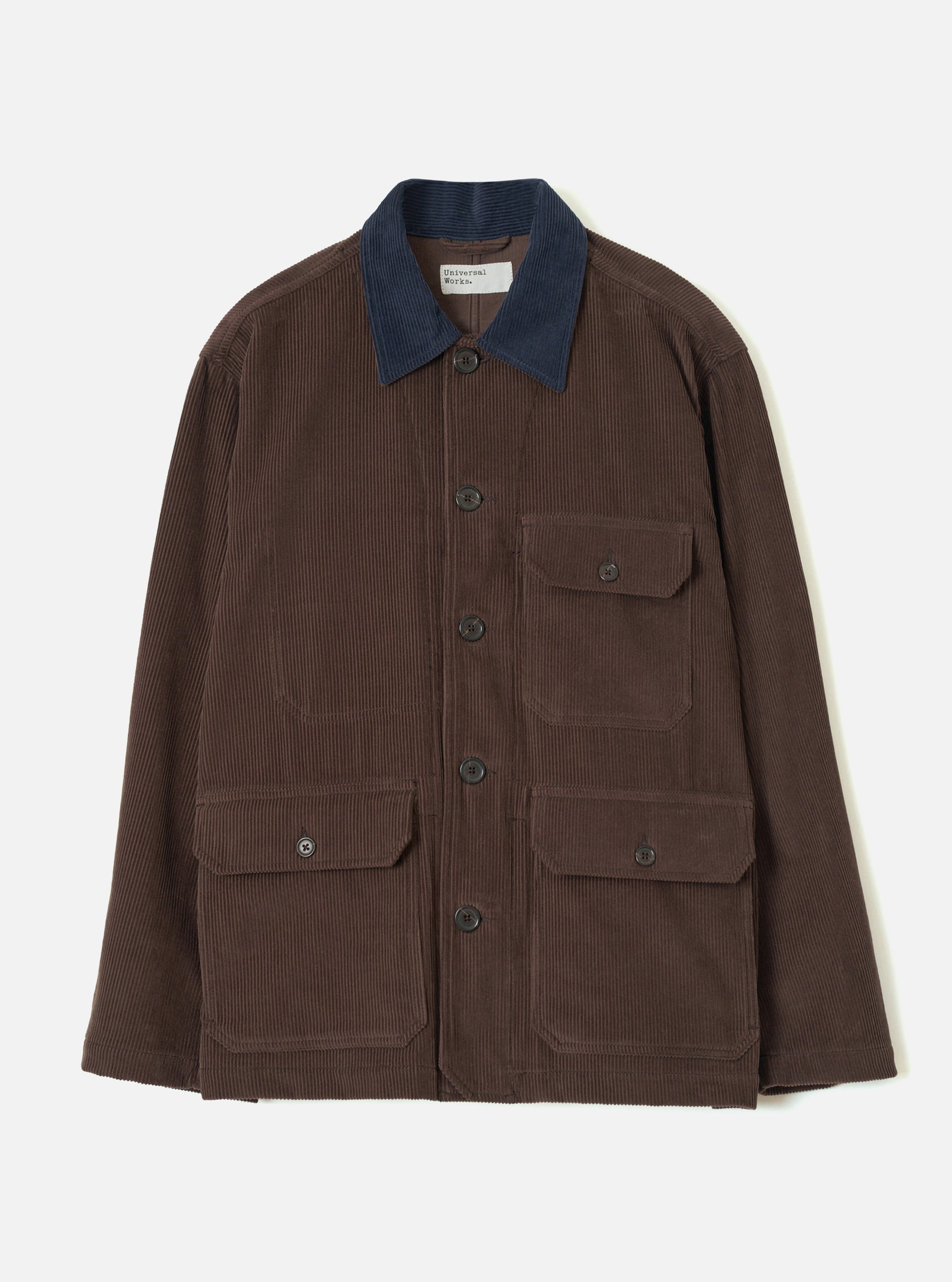 Cacao brown corduroy utility jacket laid flat on a light grey backdrop. Features a contrast navy corduroy collar, five dark buttons down the front placket, one buttoned chest patch pocket, and two large buttoned patch pockets at the hip. A Universal Works woven label sits inside the collar.