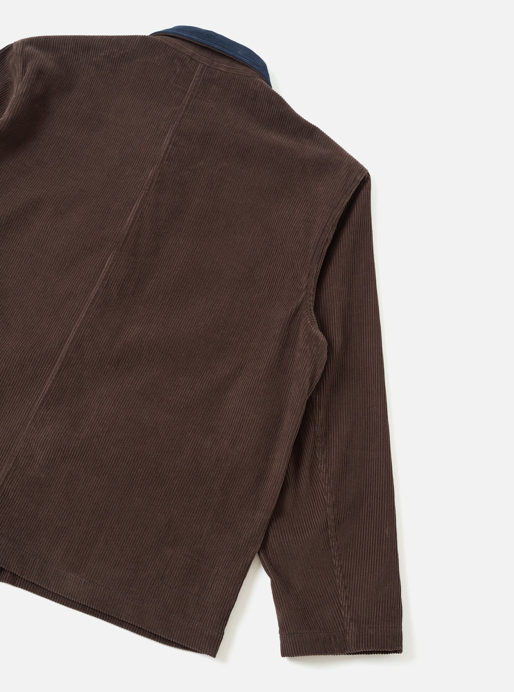 Rear and sleeve detail of a cacao brown corduroy utility jacket, shot flat on a light grey seamless backdrop. Wide-wale corduroy texture is prominent across the back panel and sleeve. A contrast navy corduroy collar is visible at the top. Diagonal back seams and a plain, unfinished hem are visible.