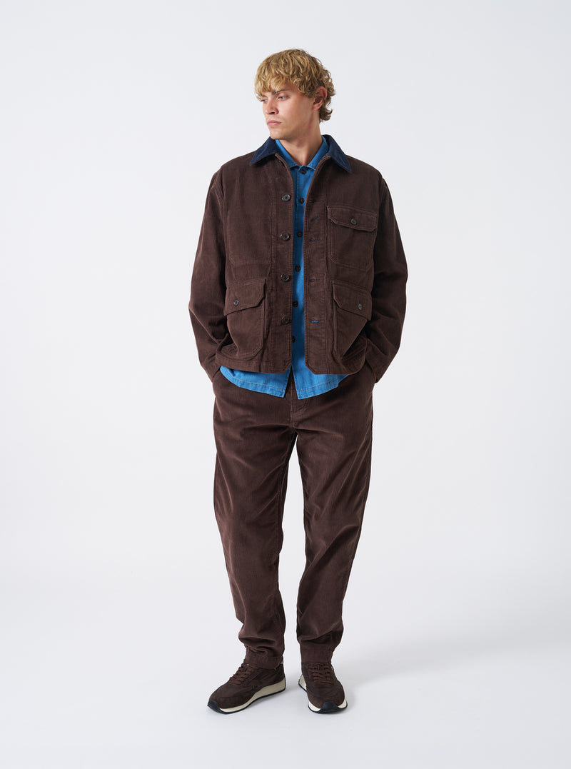 A male model wears the Cacao Cord Utility Jacket in a front-facing posture against a plain light studio background. The brown corduroy jacket features multiple utility front pockets and a neat collar.