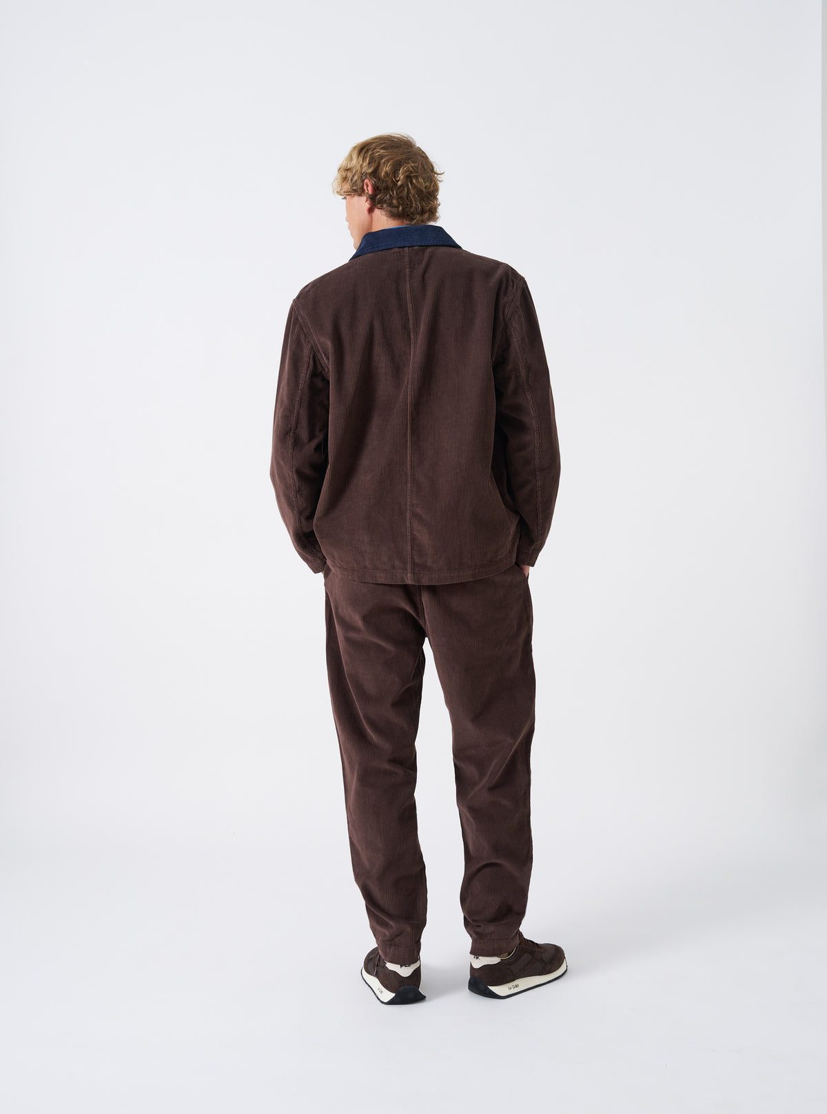A male model is wearing the Cacao Cord Utility Jacket shown from a back angled perspective against a plain studio background, emphasizing the fit, tailored shoulders, and rich cacao brown corduroy texture.