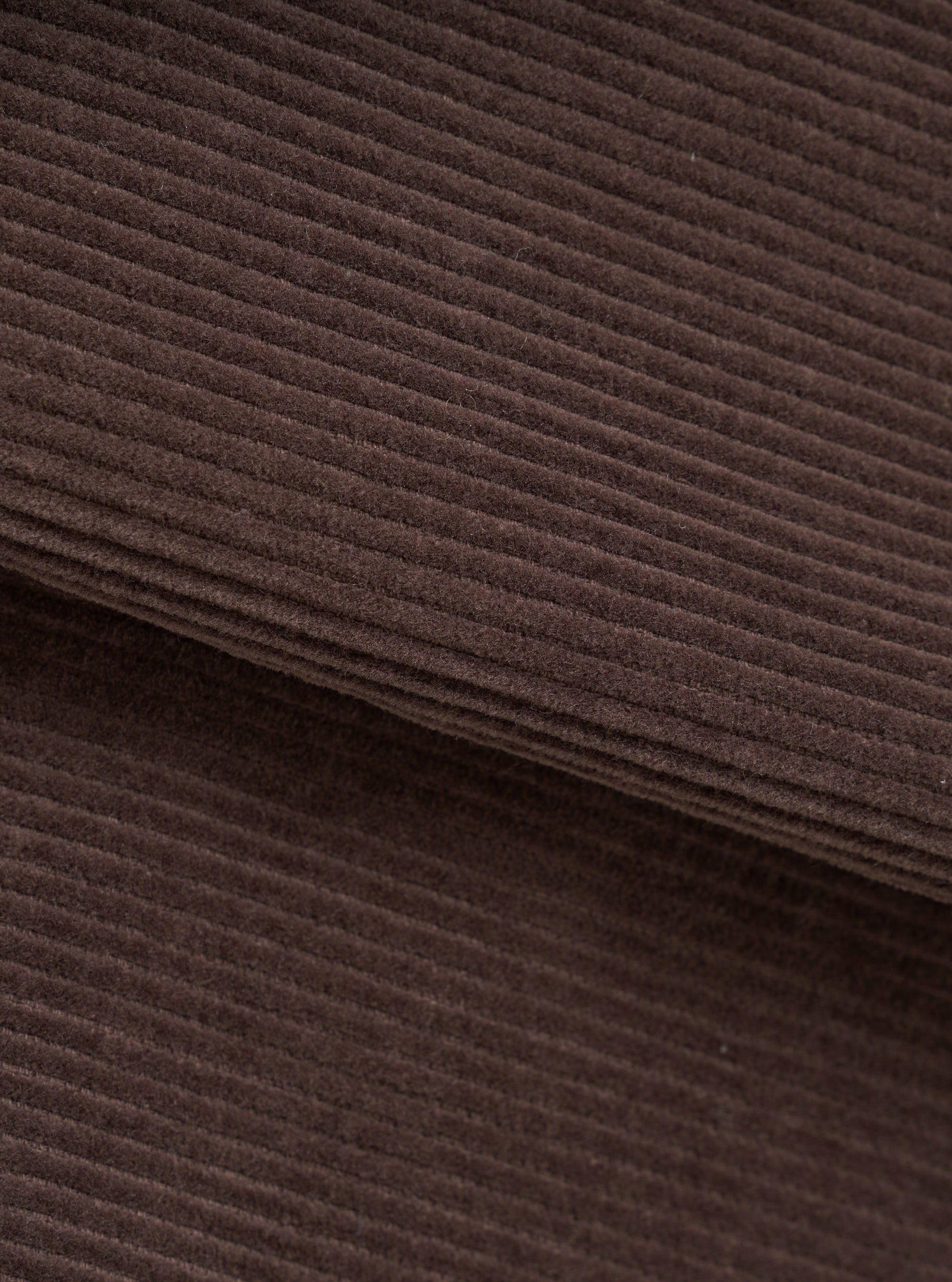 Close-up of the cacao brown corduroy fabric of the utility jacket, showing wide-wale cord texture with soft, dense pile. A folded edge runs diagonally across the frame, revealing the fabric's thickness. No background visible.