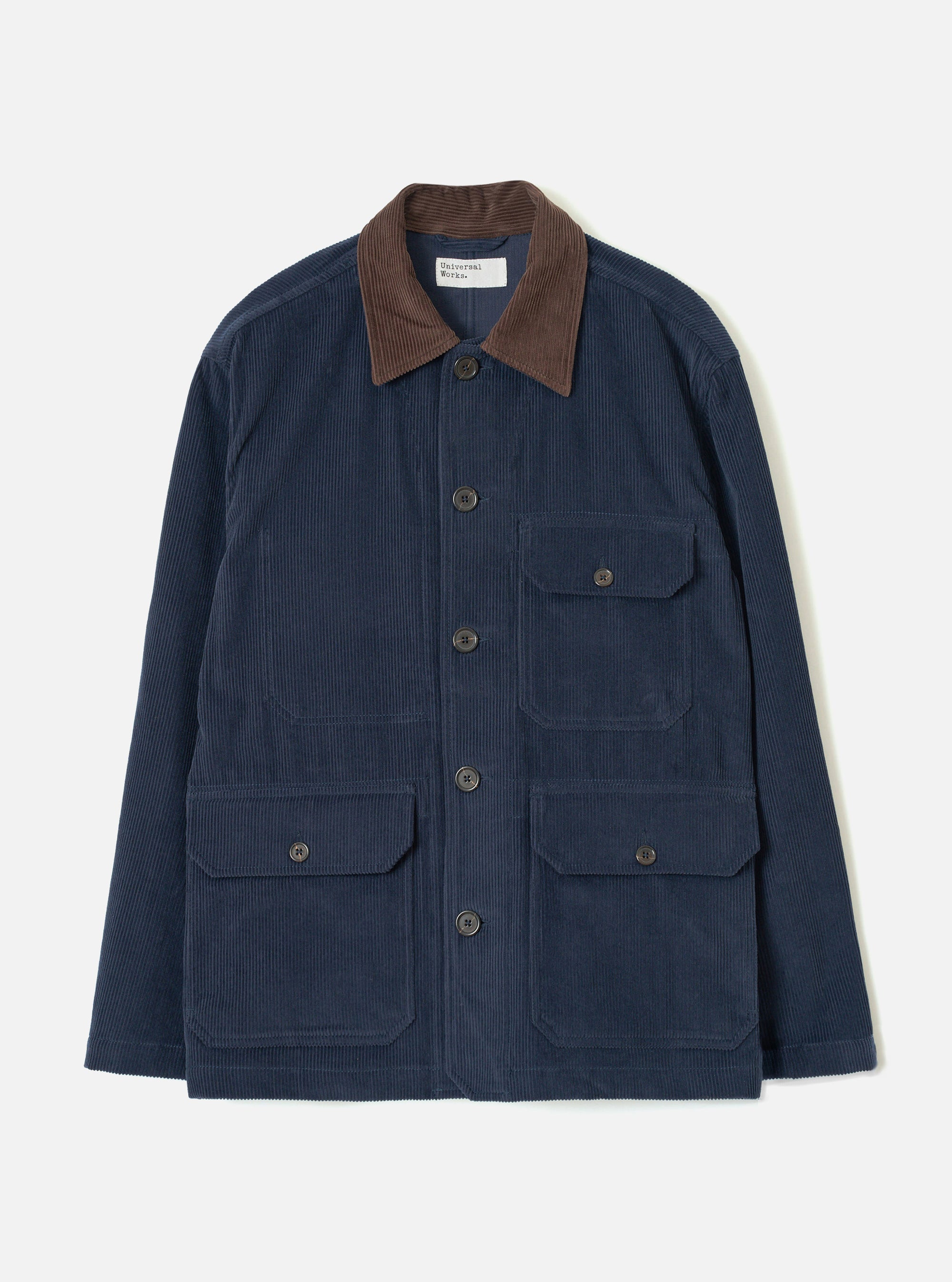 Dark navy corduroy utility jacket laid flat on a light grey studio backdrop. Features a contrast brown corduroy collar, five dark horn buttons at the front placket, two chest patch pockets with button flaps, and two larger patch pockets at the hip with button flaps. Woven brand label visible at the neck.