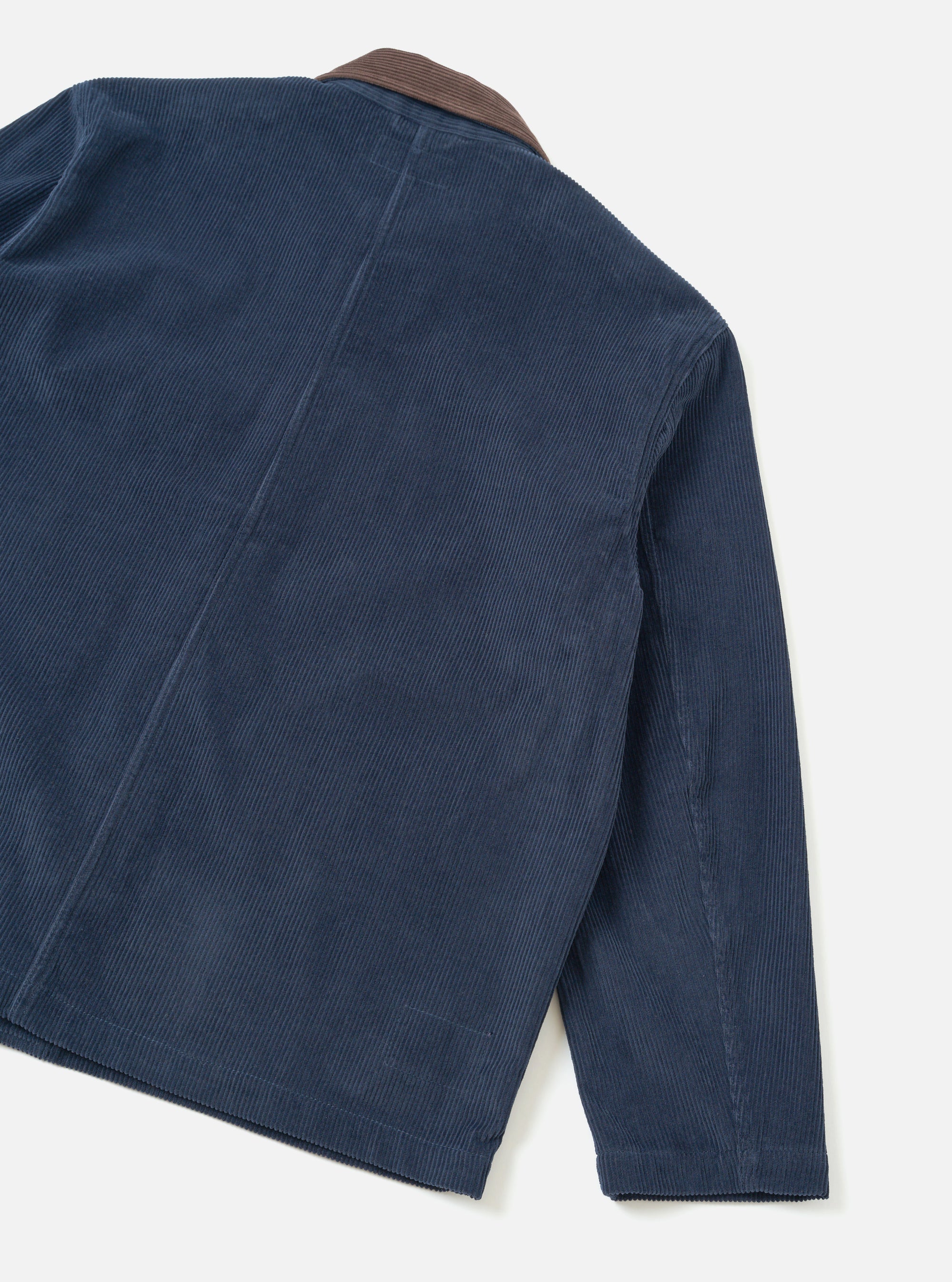 Dark navy corduroy utility jacket, flat lay on a light grey seamless backdrop. Shot shows the back and right sleeve at an angle. Wide-wale cord texture is prominent throughout. A contrast brown corduroy collar is visible at the top. Vertical back seams and a straight hem with visible stitching are shown.