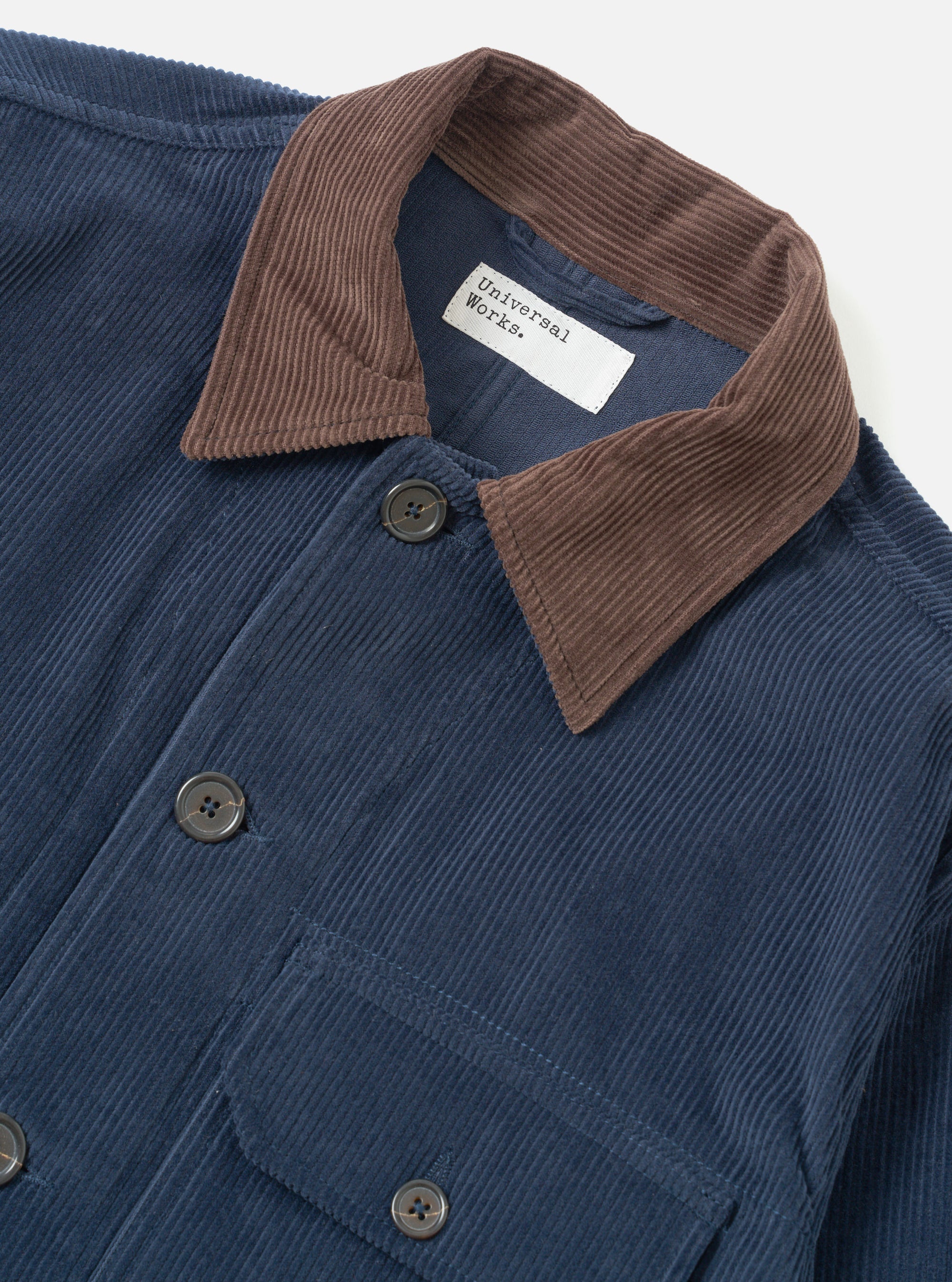 Close-up of a Universal Works dark navy corduroy utility jacket on a plain white backdrop. The wide-wale cord body contrasts with a brown corduroy collar. Dark four-hole buttons fasten the front placket. A chest patch pocket is visible at lower left. The woven brand label sits inside the open collar.