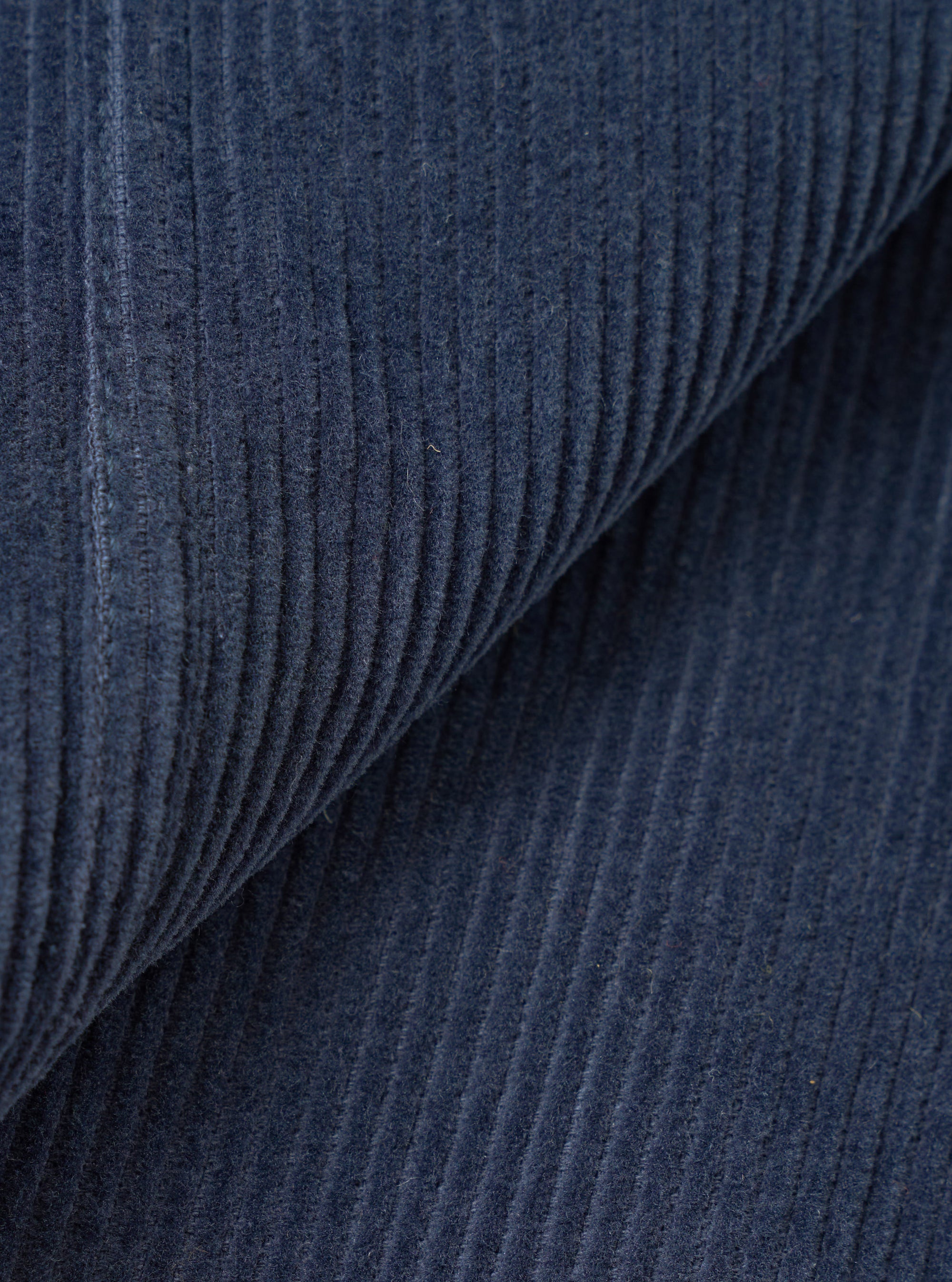 Close-up of dark navy corduroy fabric, showing wide-wale cord texture in detail. The fabric is folded to reveal the raised ribs and soft pile. A seam is visible on the left edge. The shot fills the entire frame with no background visible.