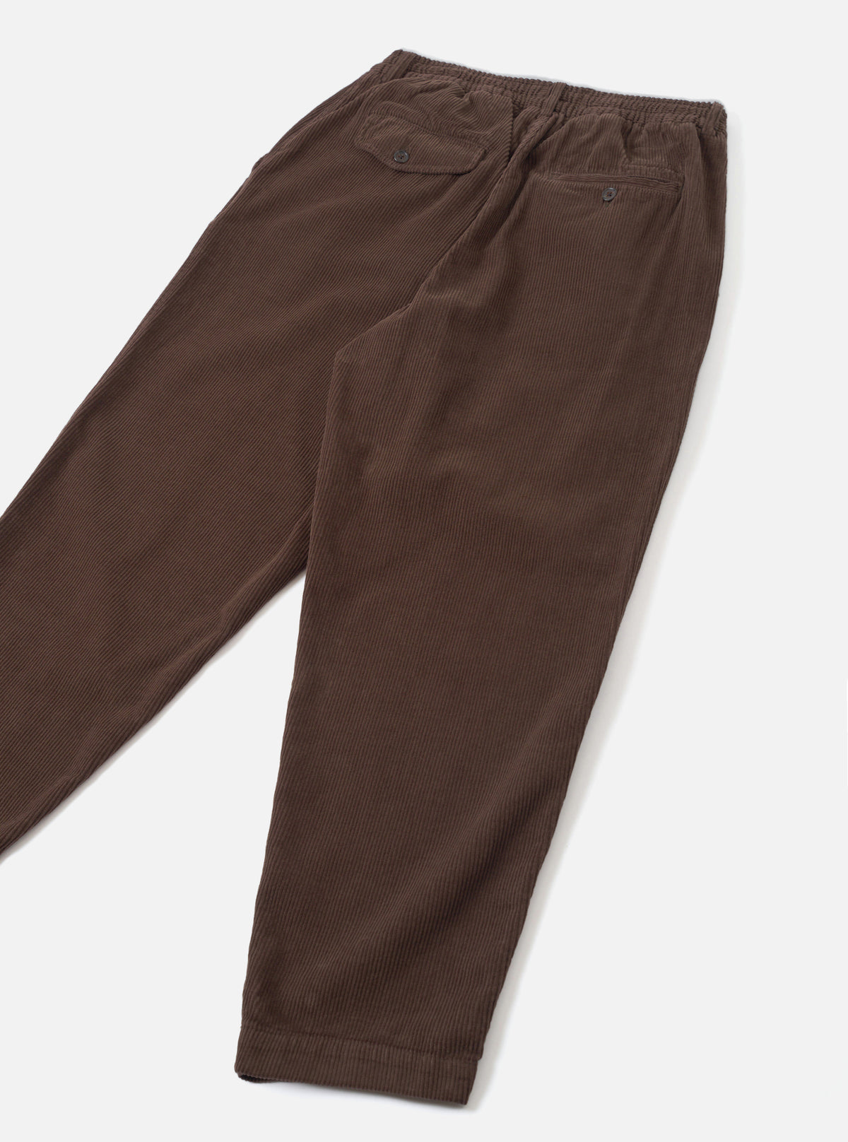 A full rear shot of the Cacao Cord Pleated Track Pant laid flat against a clean white background. The image highlights the back welt pockets, elasticated waistband seam, and rich brown corduroy fabric structure.