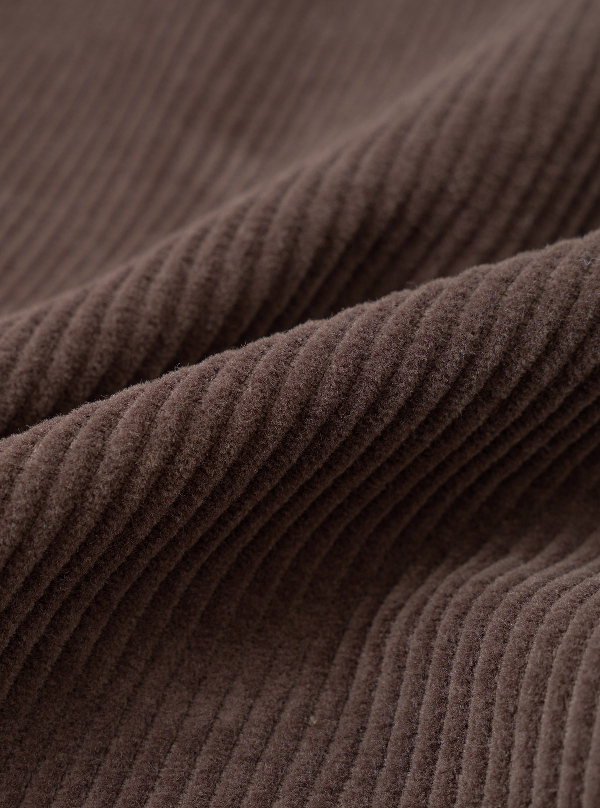 A detailed close-up fabric swatch of the Cacao Cord Pleated Track Pant presented on a stark white background, highlighting the distinct fine-wale ridges of the rich cacao brown cotton corduroy fabric.