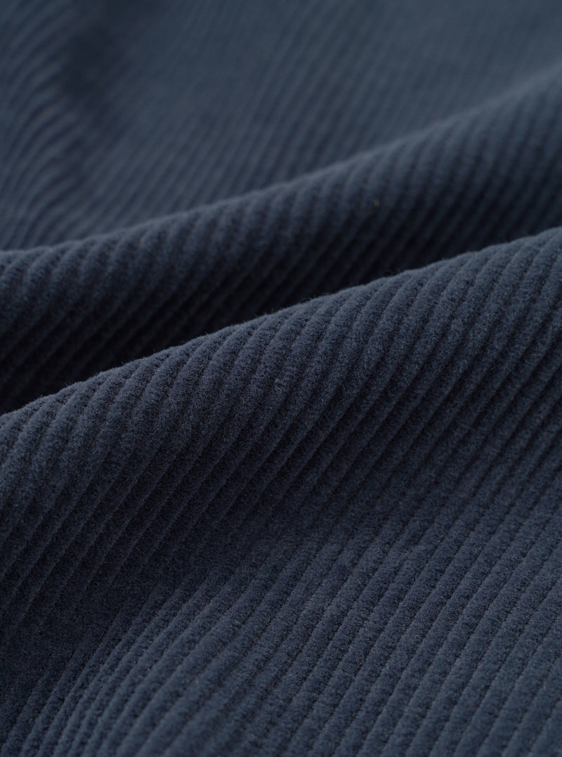 A macro fabric swatch photo of the Dark Navy Cord Pleated Track Pant set against a plain white background, highlighting the soft texture and fine ribbed lines of the navy blue cotton corduroy material.